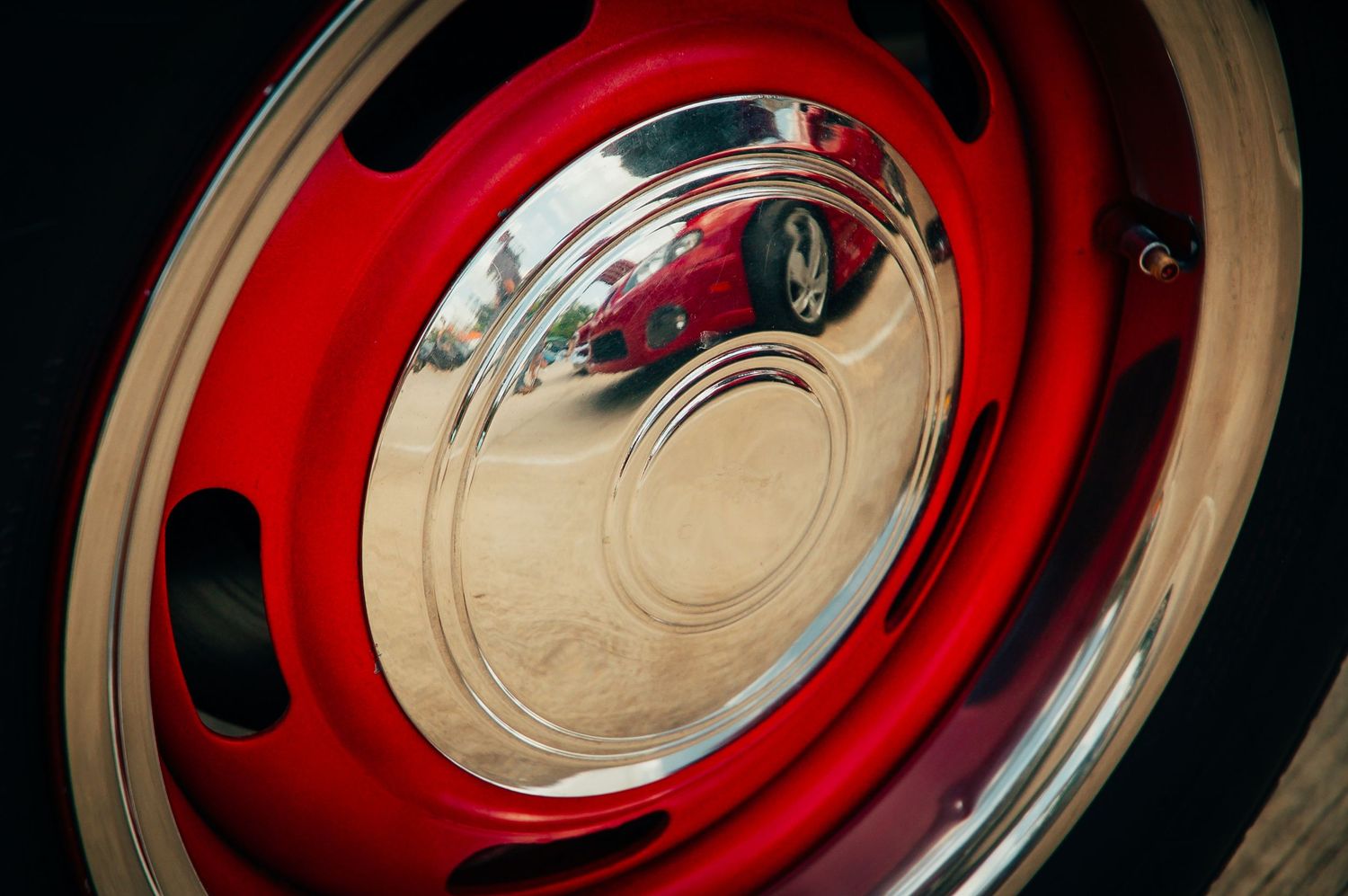 Shiny red classic car wheel with chrome hubcap reflecting a red vehicle at a car show.