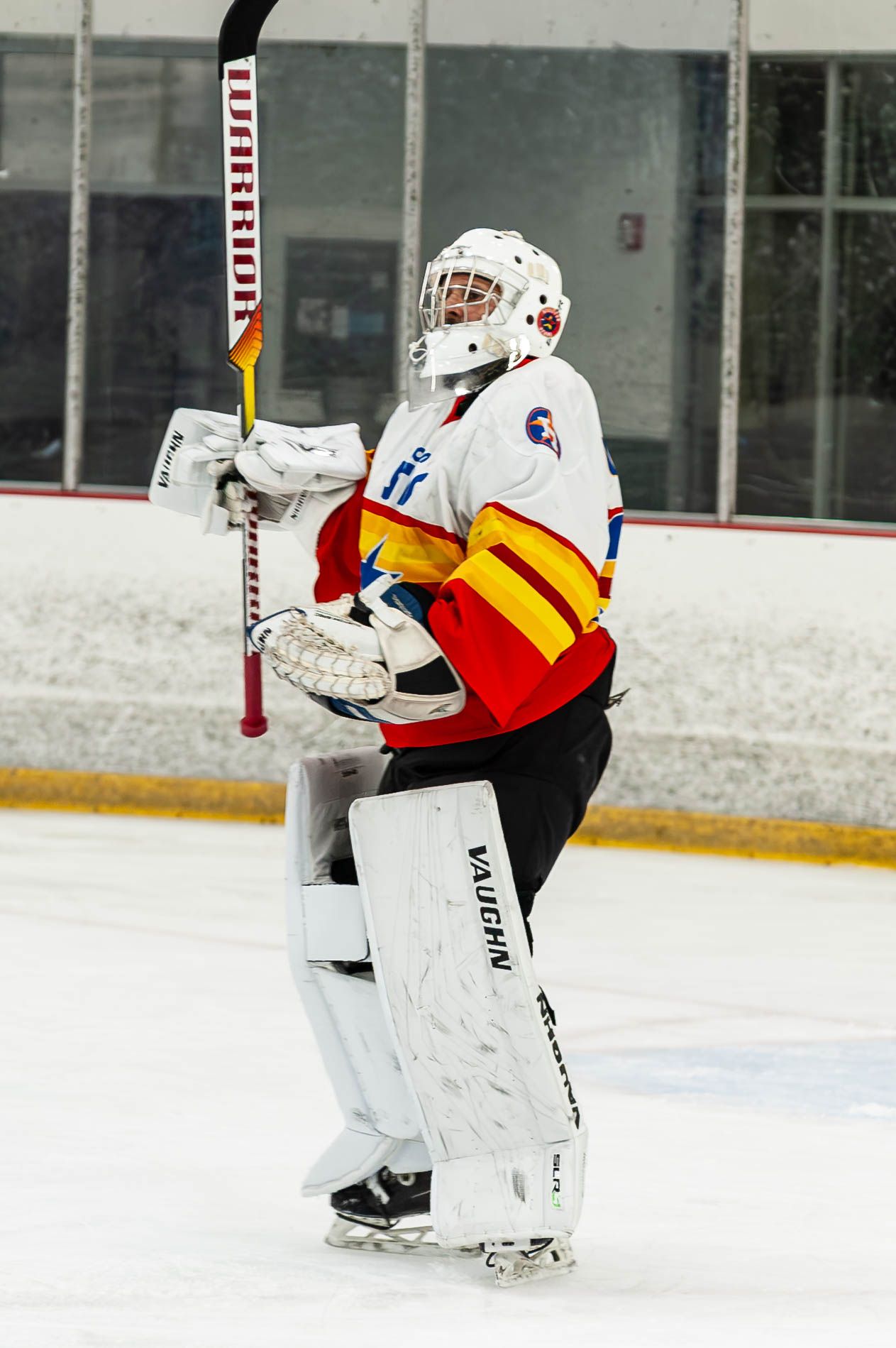 A hockey goalie in white and red Vaughn gear holds a Warrior stick on an indoor ice rink.