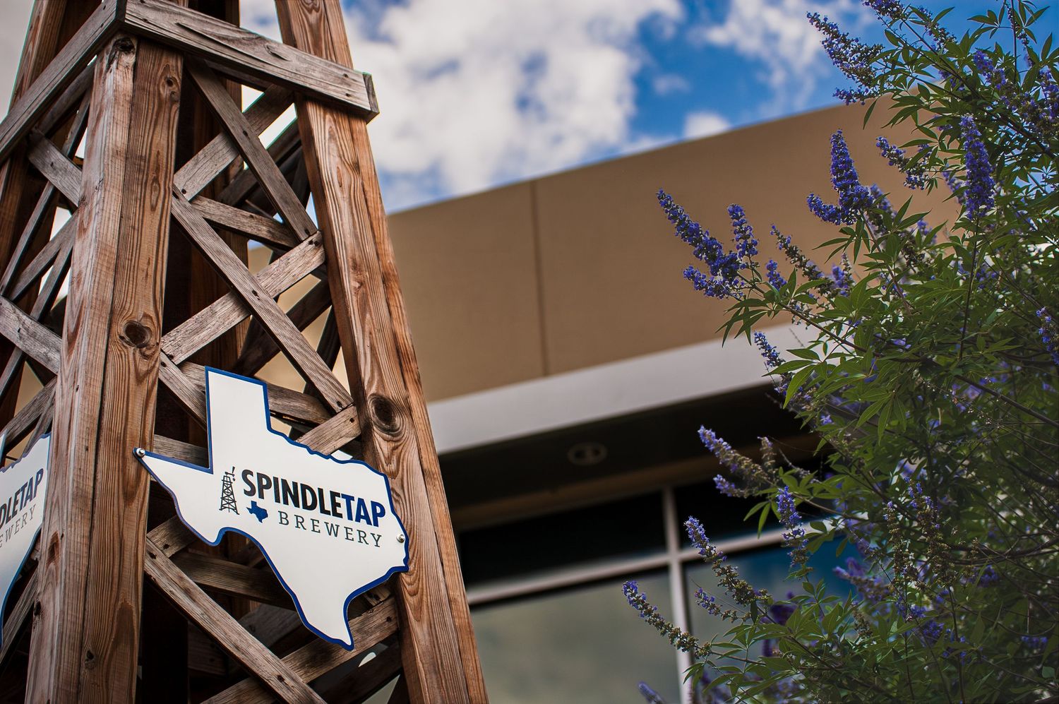 Spindletap Brewery sign shaped like Texas on a wooden structure with purple flowers and blue sky.