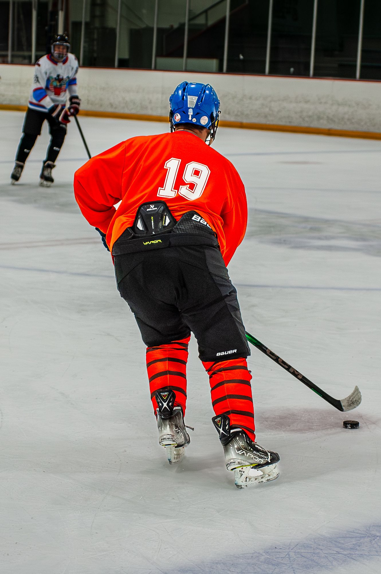 Hockey player wearing number 19 red jersey skating with puck on indoor ice rink.