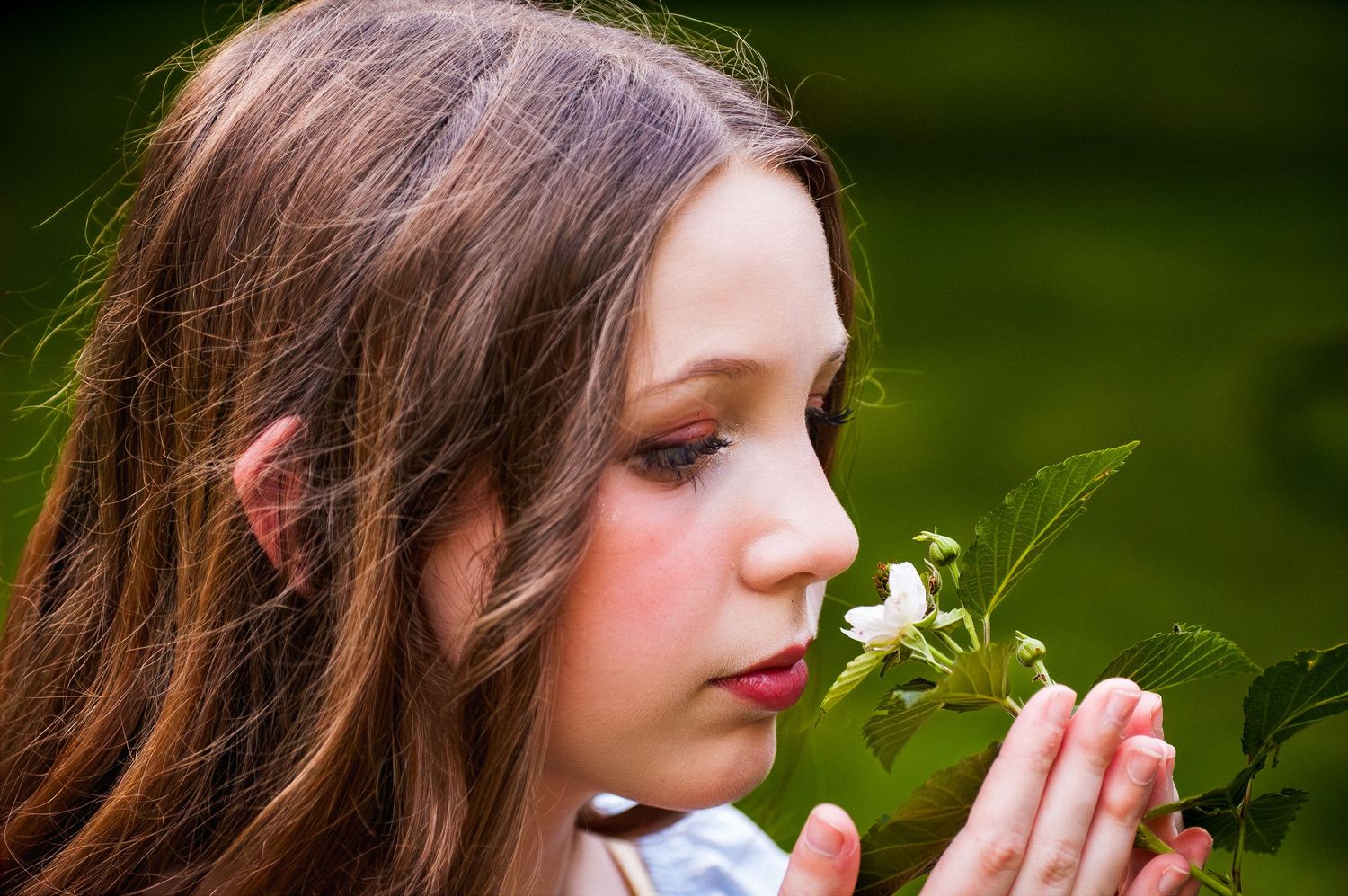 Young girl with brown hair smelling a delicate white flower outdoors in natural light.