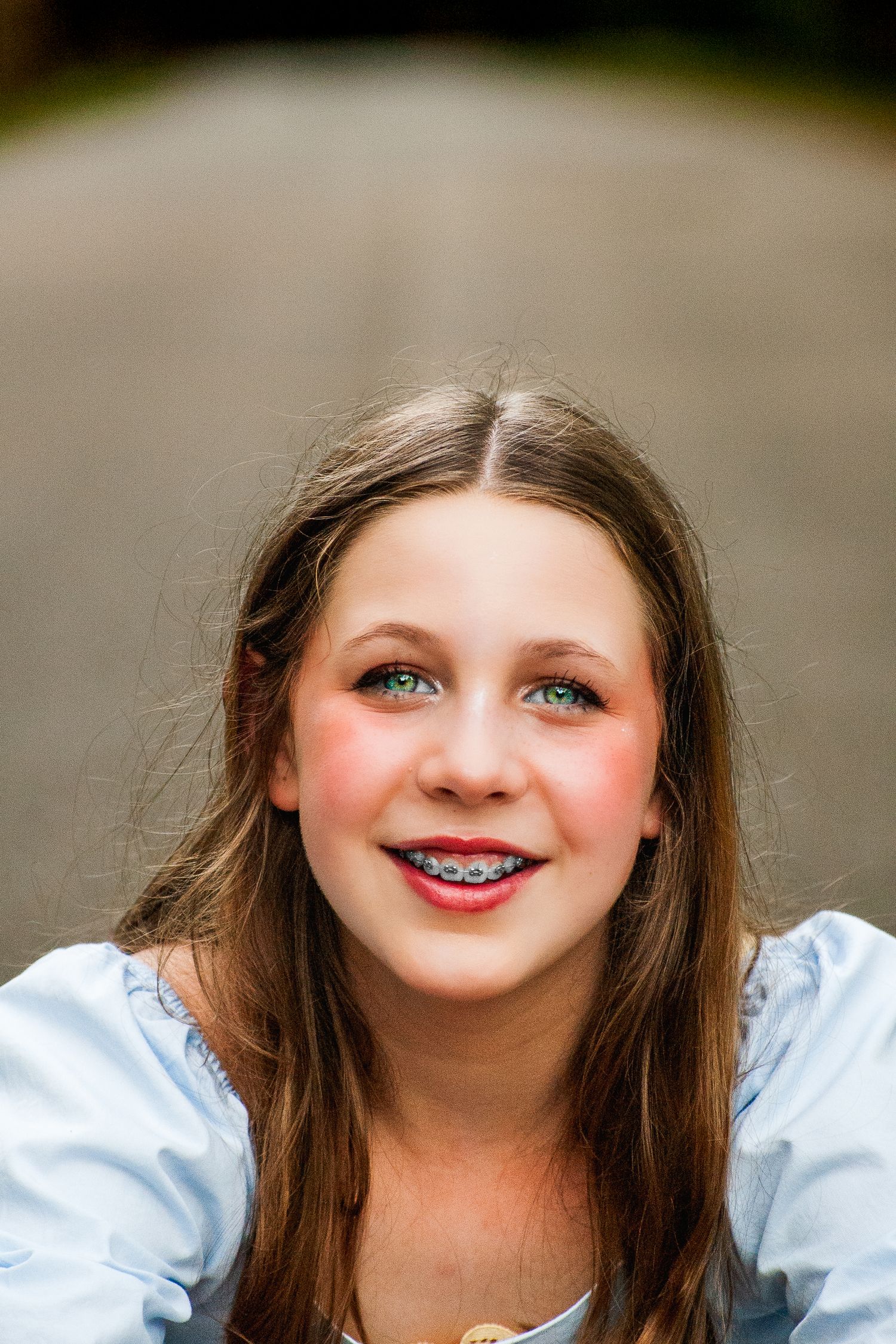 Smiling teenage girl with brown hair, green eyes, and braces wearing a light blue top outdoors.