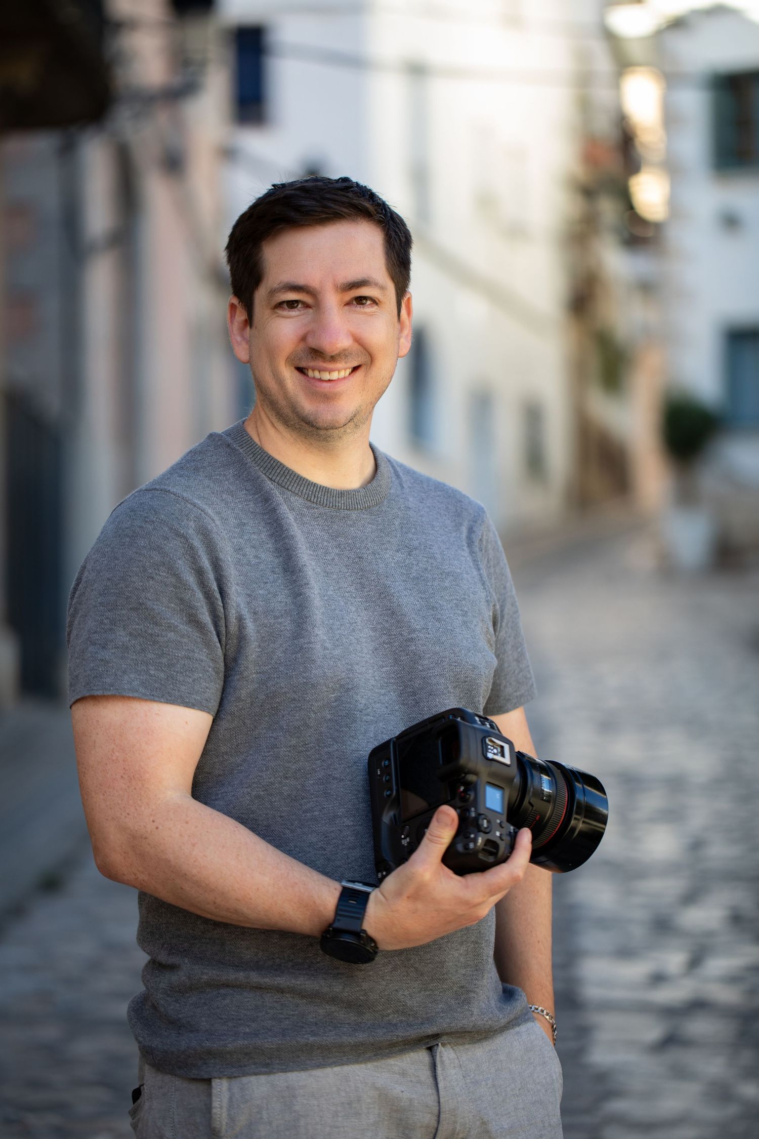 Smiling man holding a DSLR camera on a cobblestone street wearing a gray t-shirt.