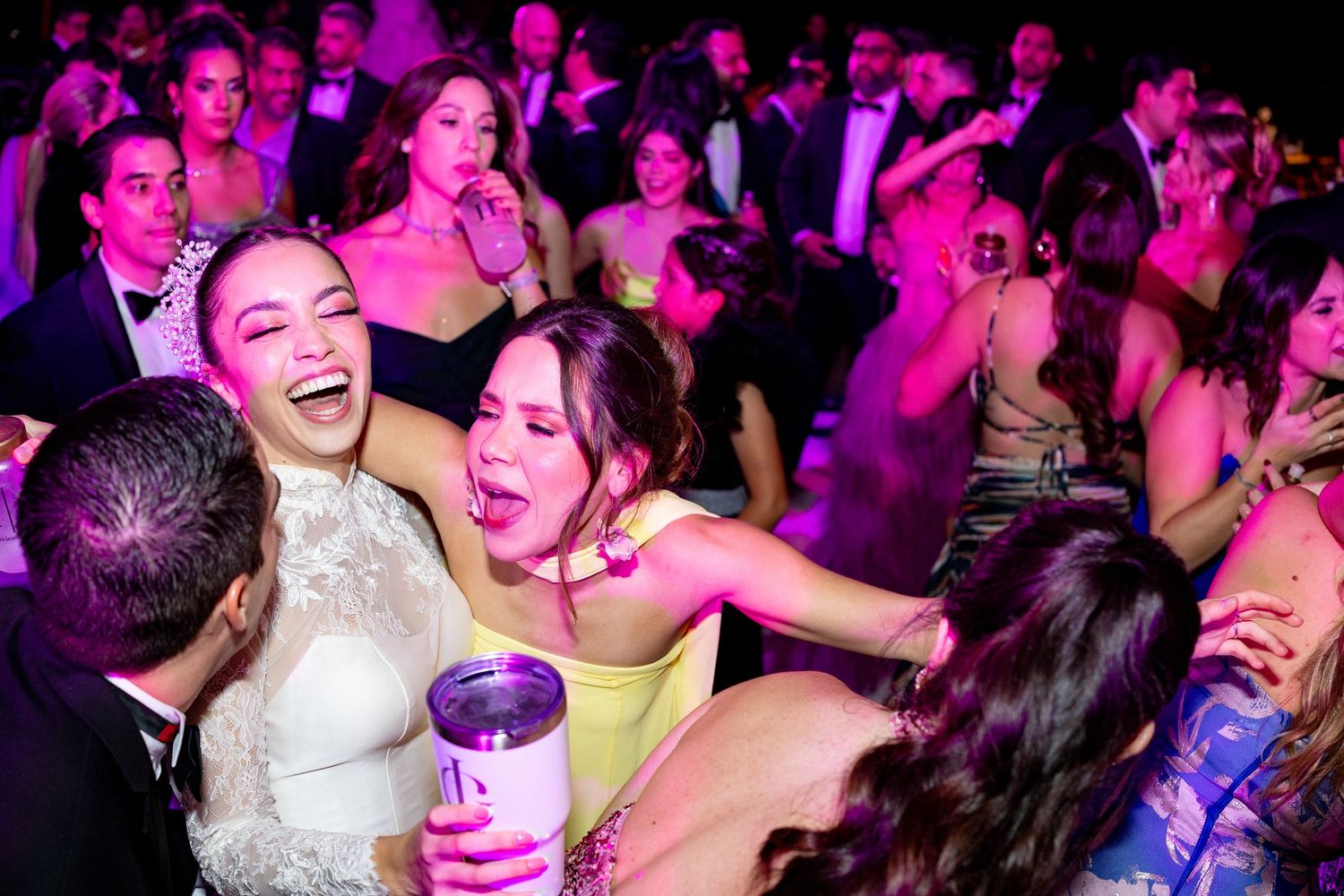 Joyful bride in white lace gown laughing with guests in yellow and black dresses at wedding reception.