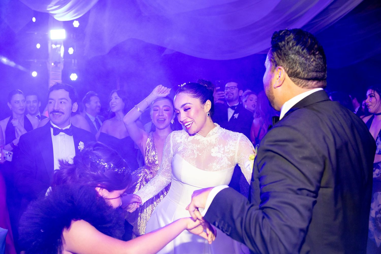 Bride in lace gown dancing joyfully with guests under purple lights at vibrant wedding reception.