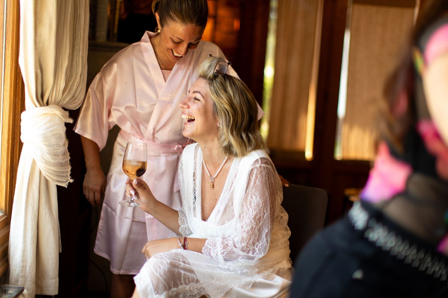 Joyful bride in lace robe laughing while holding champagne glass during wedding preparations.