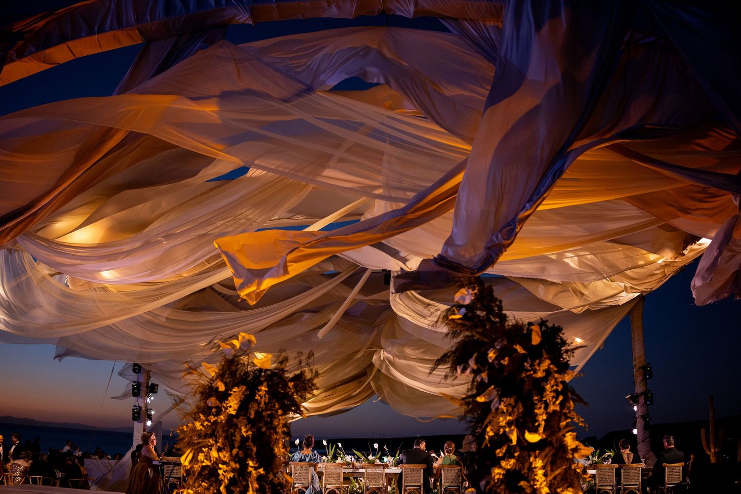 Dramatic evening wedding canopy with golden-lit billowing fabric drapes and floral arrangements at dusk.