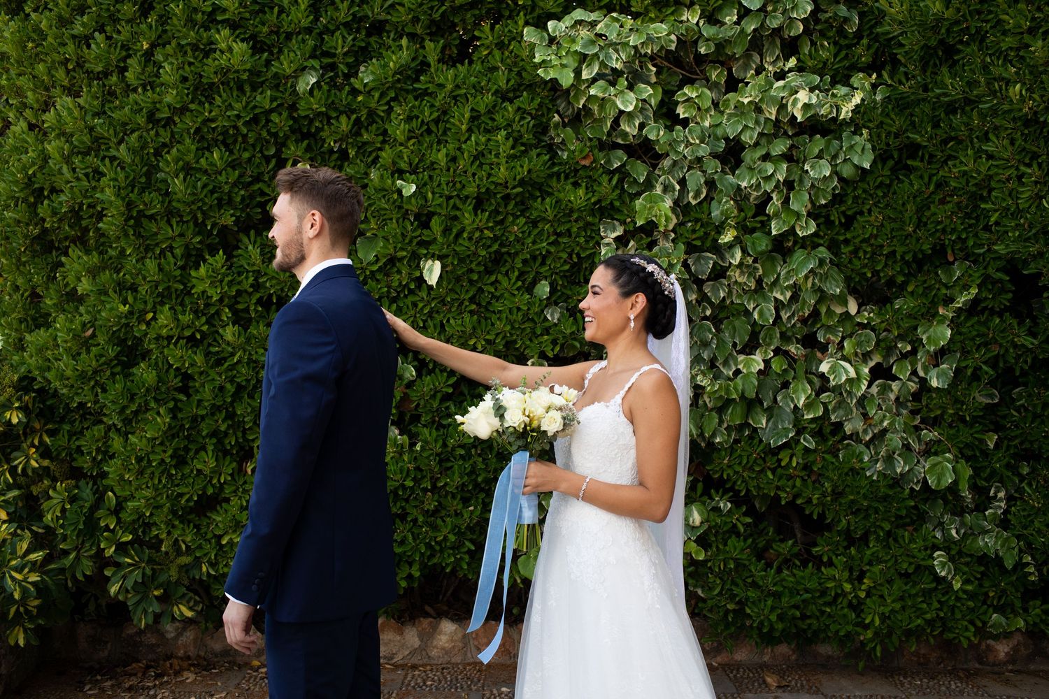 Bride taps groom on shoulder during emotional first look moment in front of green hedge wall.