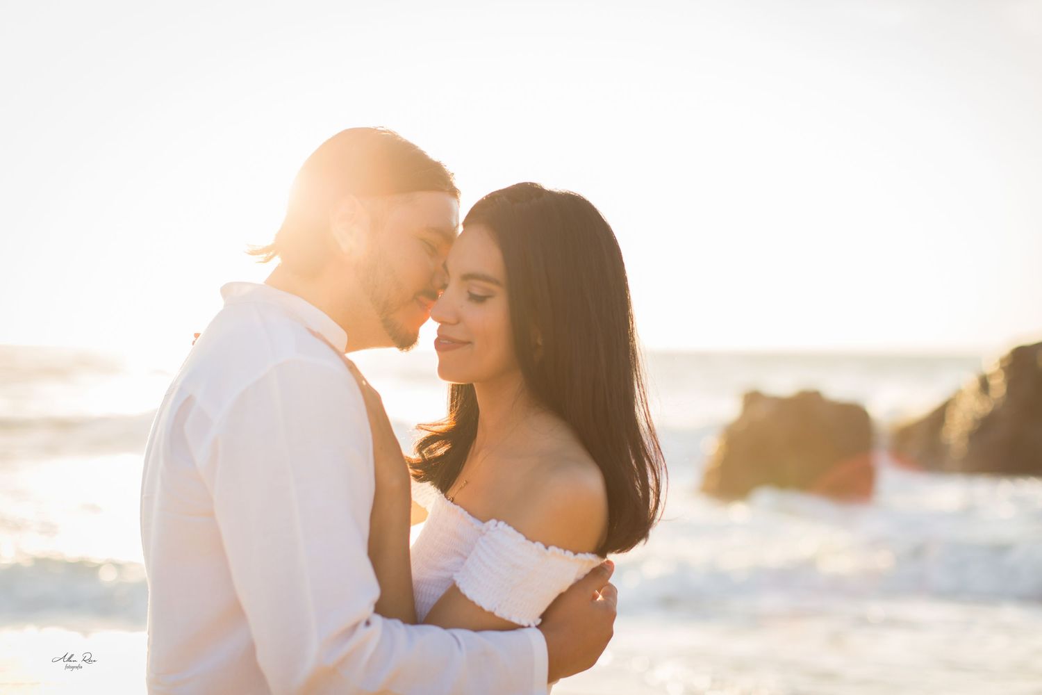 Couple embraces romantically at sunset on a beach, golden light glowing behind them near ocean waves.