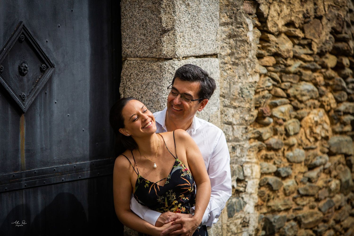 Couple embracing tenderly against a rustic stone wall and vintage wooden door in a charming European setting.