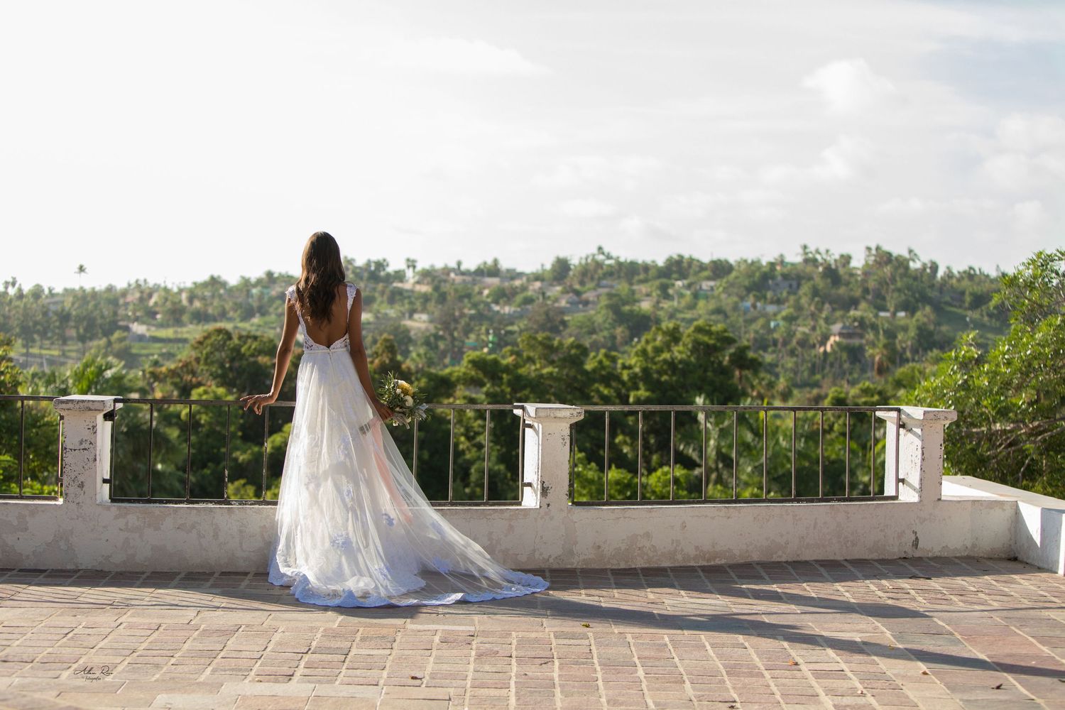 Bride in white gown with long train stands at railing overlooking tropical treetop panorama.