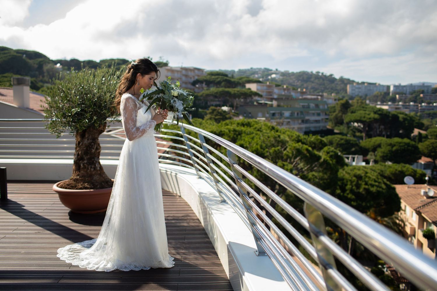 Bride in a flowing white lace wedding dress holds a bouquet while standing on a terrace with a scenic hillside view.