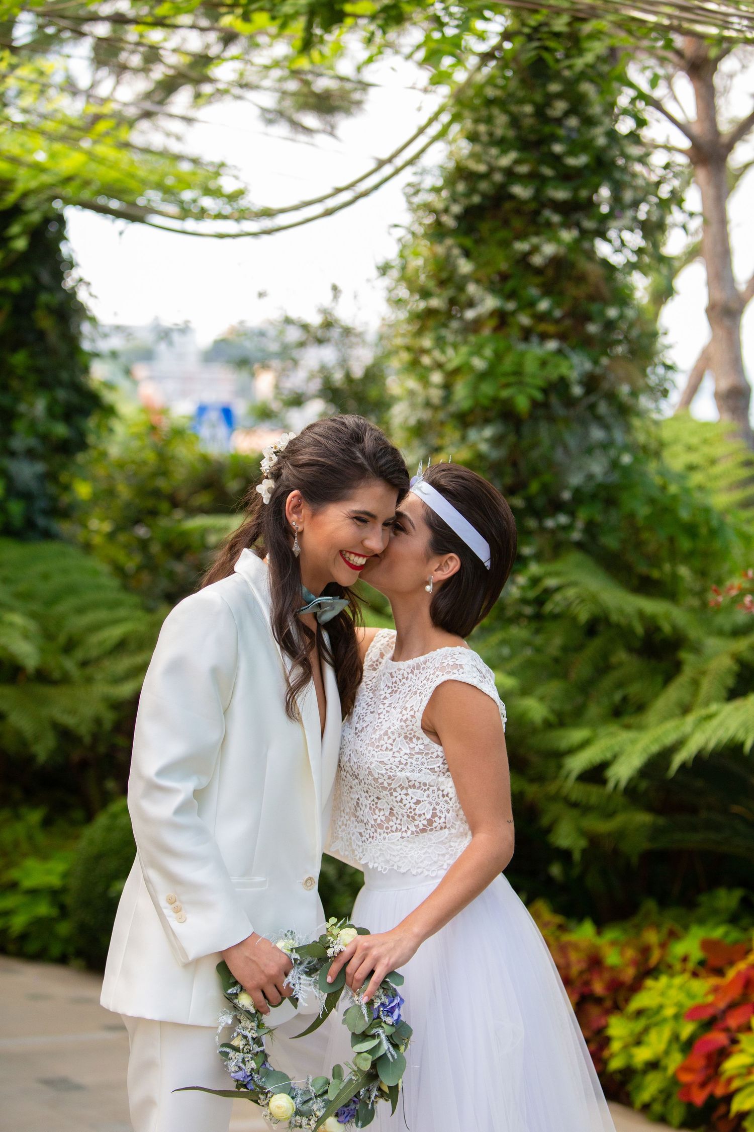 Two brides share a joyful kiss in a garden, one in a white suit and one in a lace wedding dress with headband.
