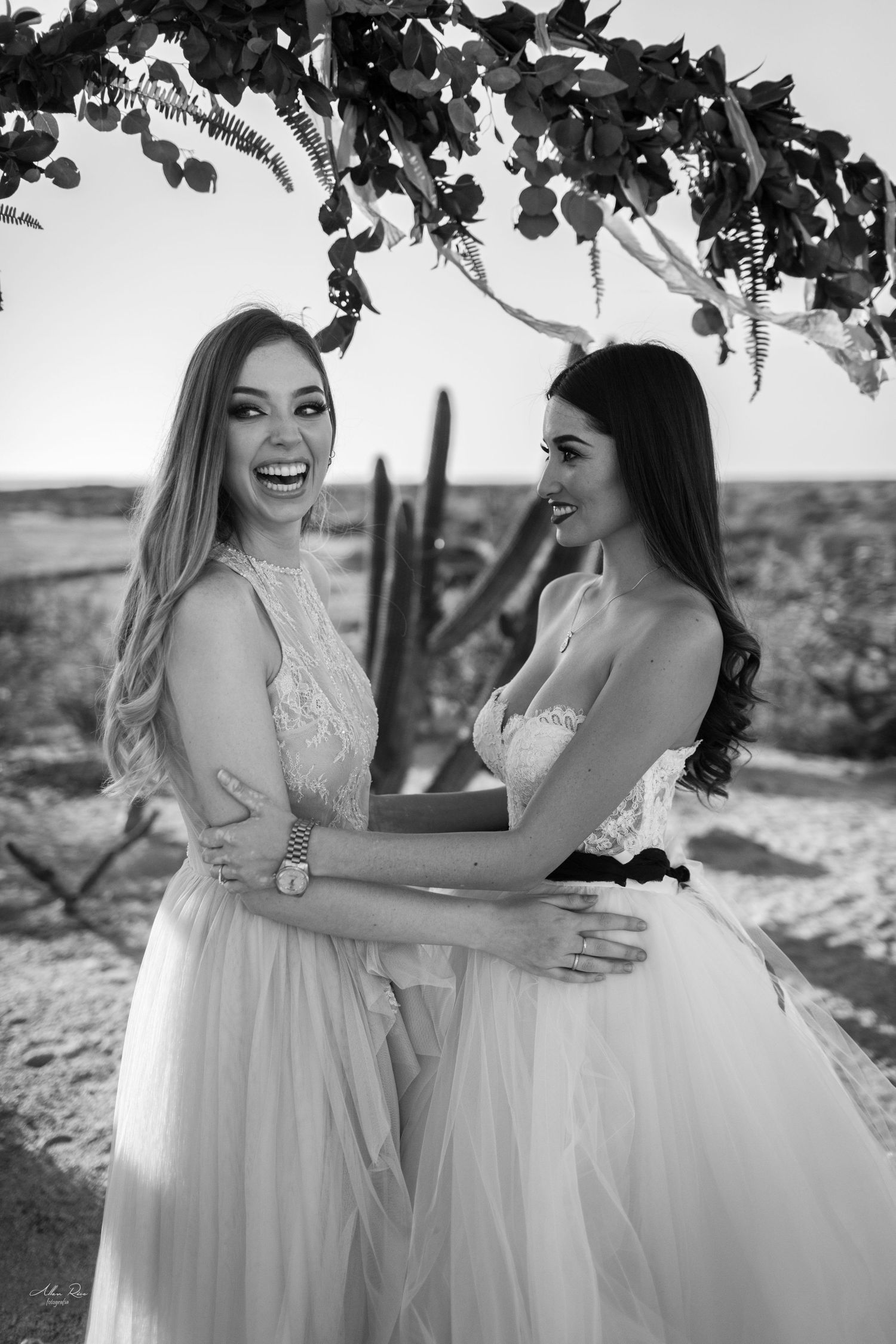 Black and white photo of two laughing brides embracing under a floral arch in the desert.