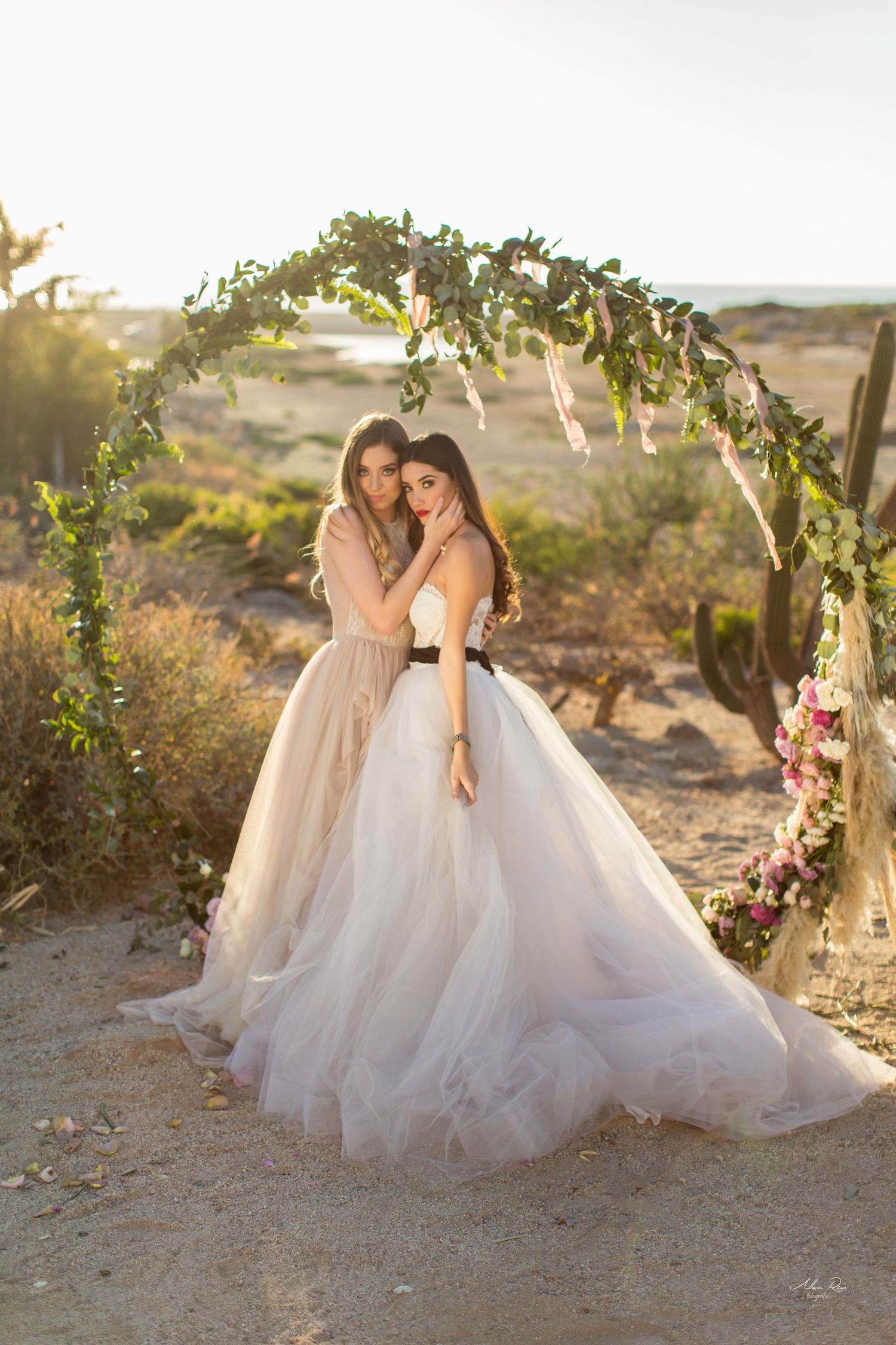 Two brides in flowing tulle gowns embrace under a floral arch in a desert wedding setting at golden hour.
