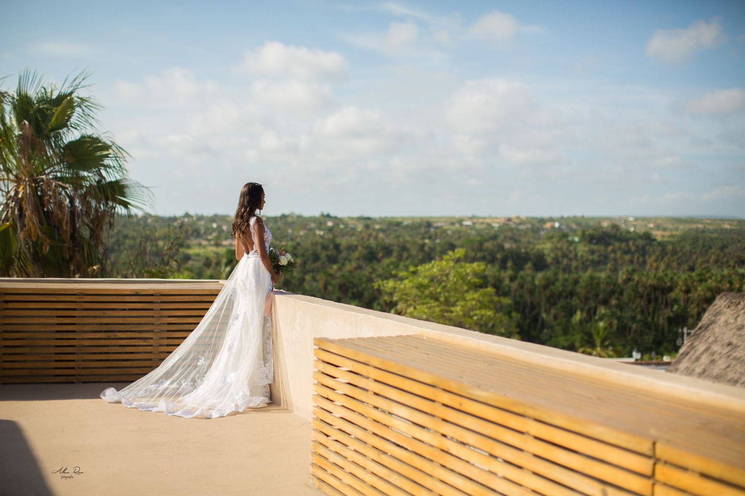 Bride in white gown with lace train stands on rooftop overlooking lush tropical jungle landscape.