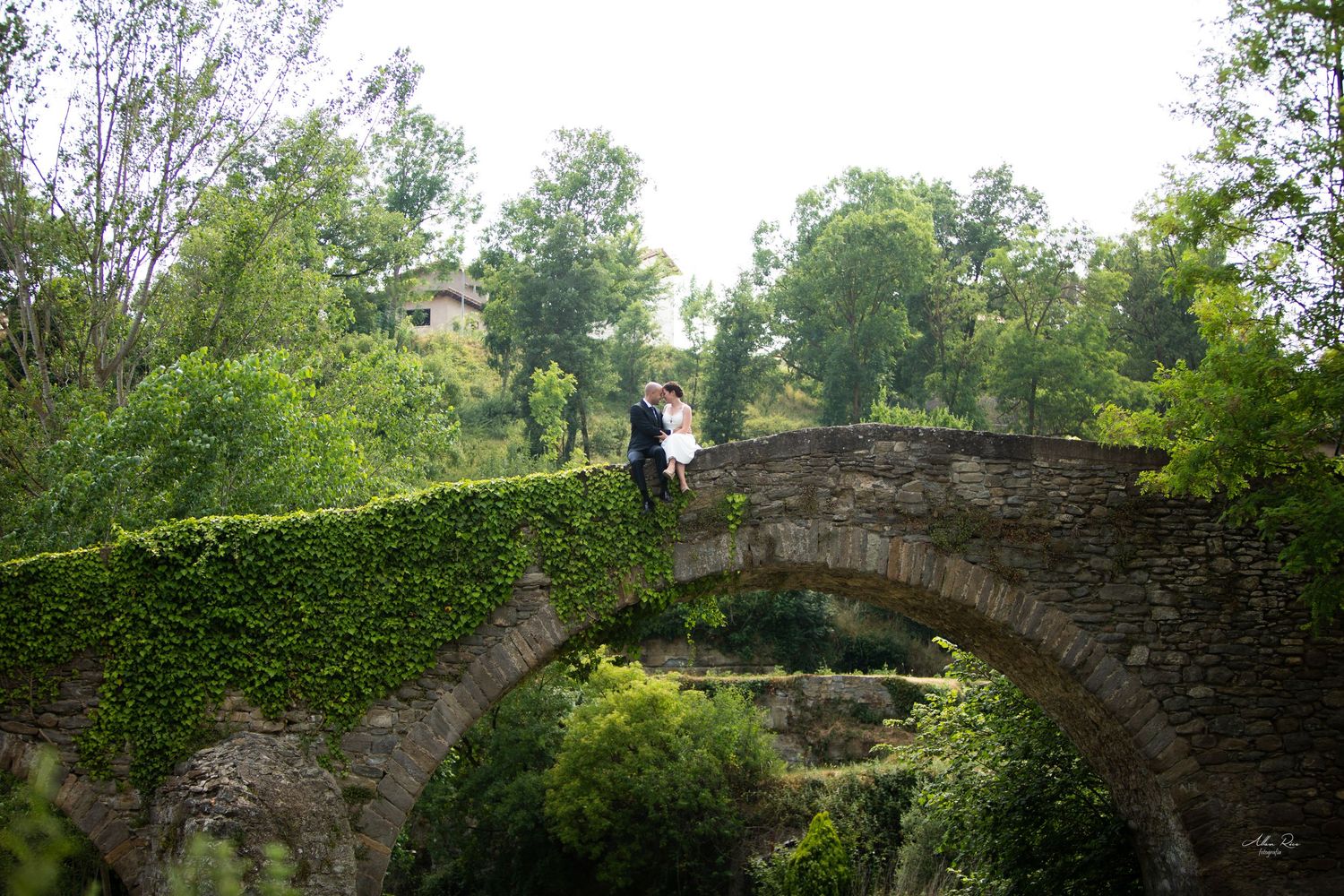 Bride and groom standing on an ancient ivy-covered stone bridge arch surrounded by lush greenery.
