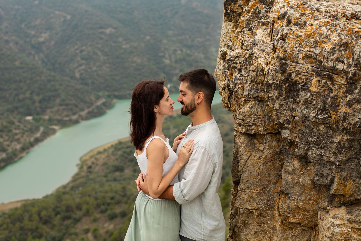 Couple embracing intimately against a rugged stone wall with a scenic river valley visible below.
