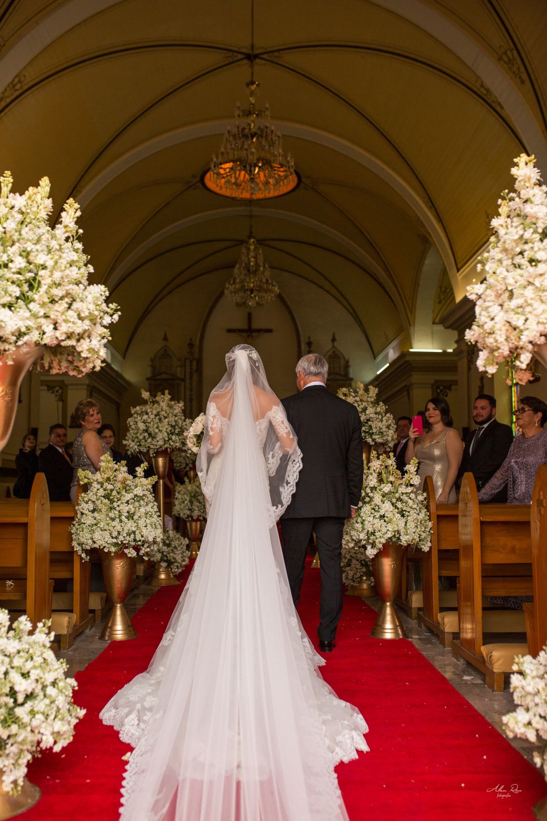 Bride in lace gown with long veil walks down red carpet aisle of ornate church with father.