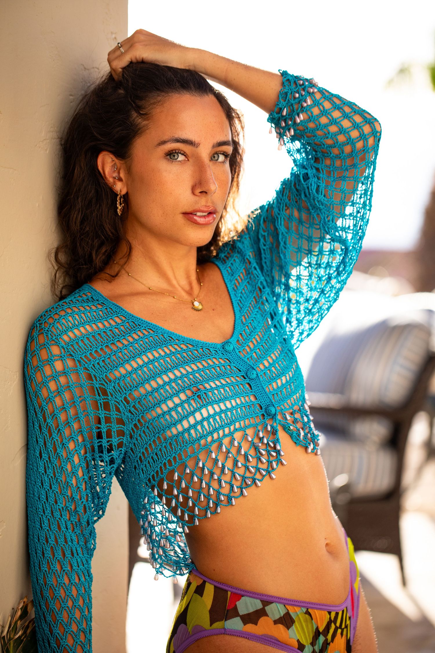 Woman in turquoise crochet mesh top and colorful skirt posing against a sunlit white wall outdoors.