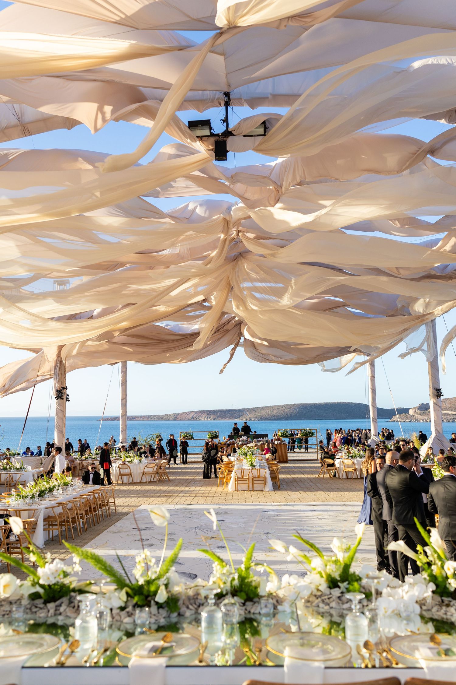Elegant beachside wedding reception under flowing white fabric ceiling with floral centerpieces and ocean views.