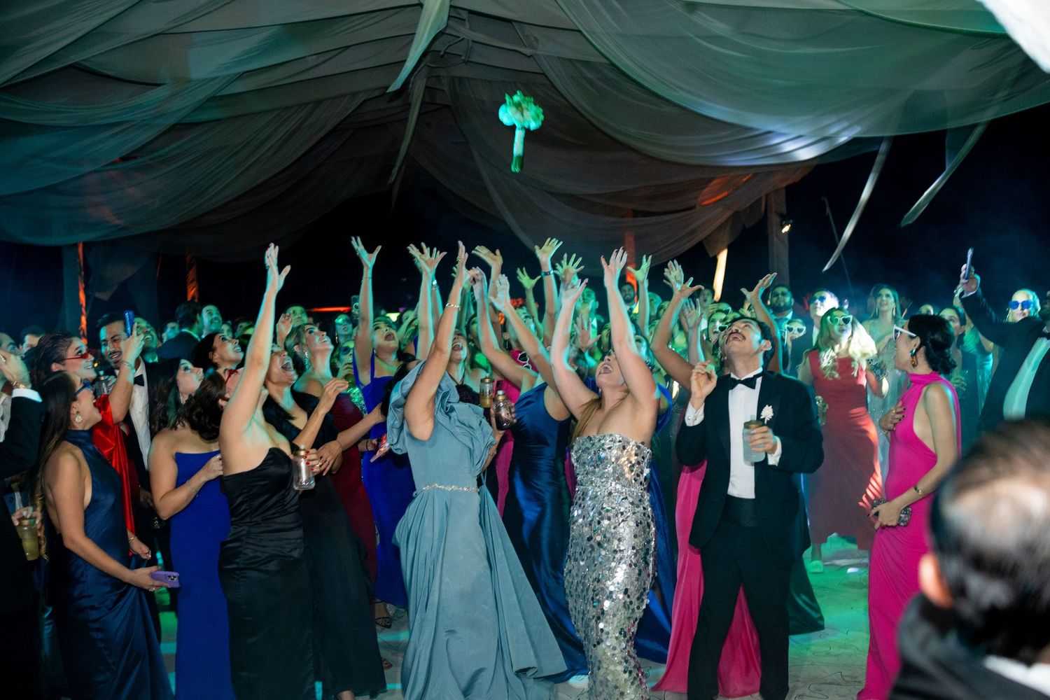 Wedding guests in formal attire raise hands together on a teal-lit dance floor under a draped tent.
