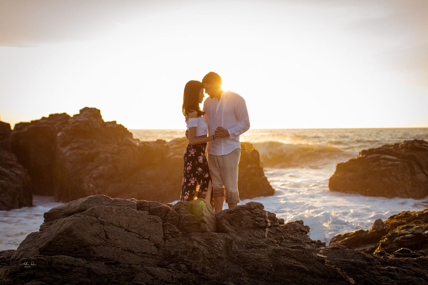 Couple standing close together on rocky coastline during stunning golden sunset.