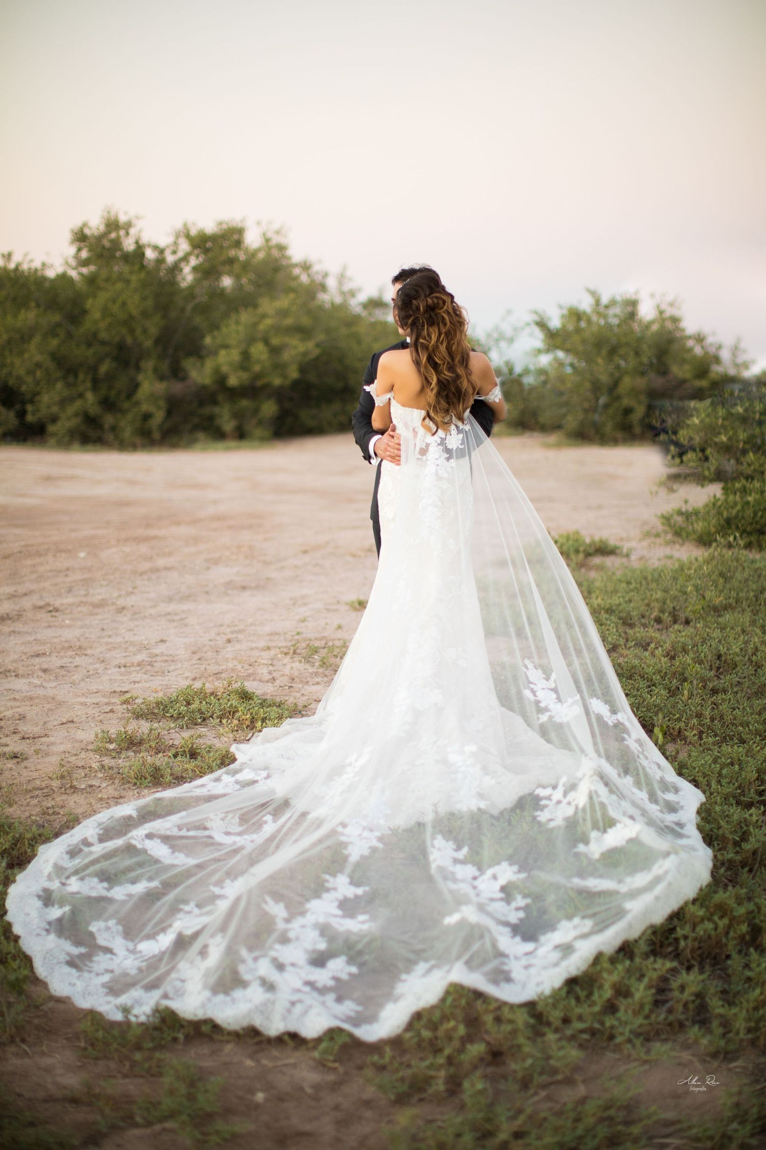Bride in lace wedding gown with long cathedral veil embraced by groom in outdoor desert setting.