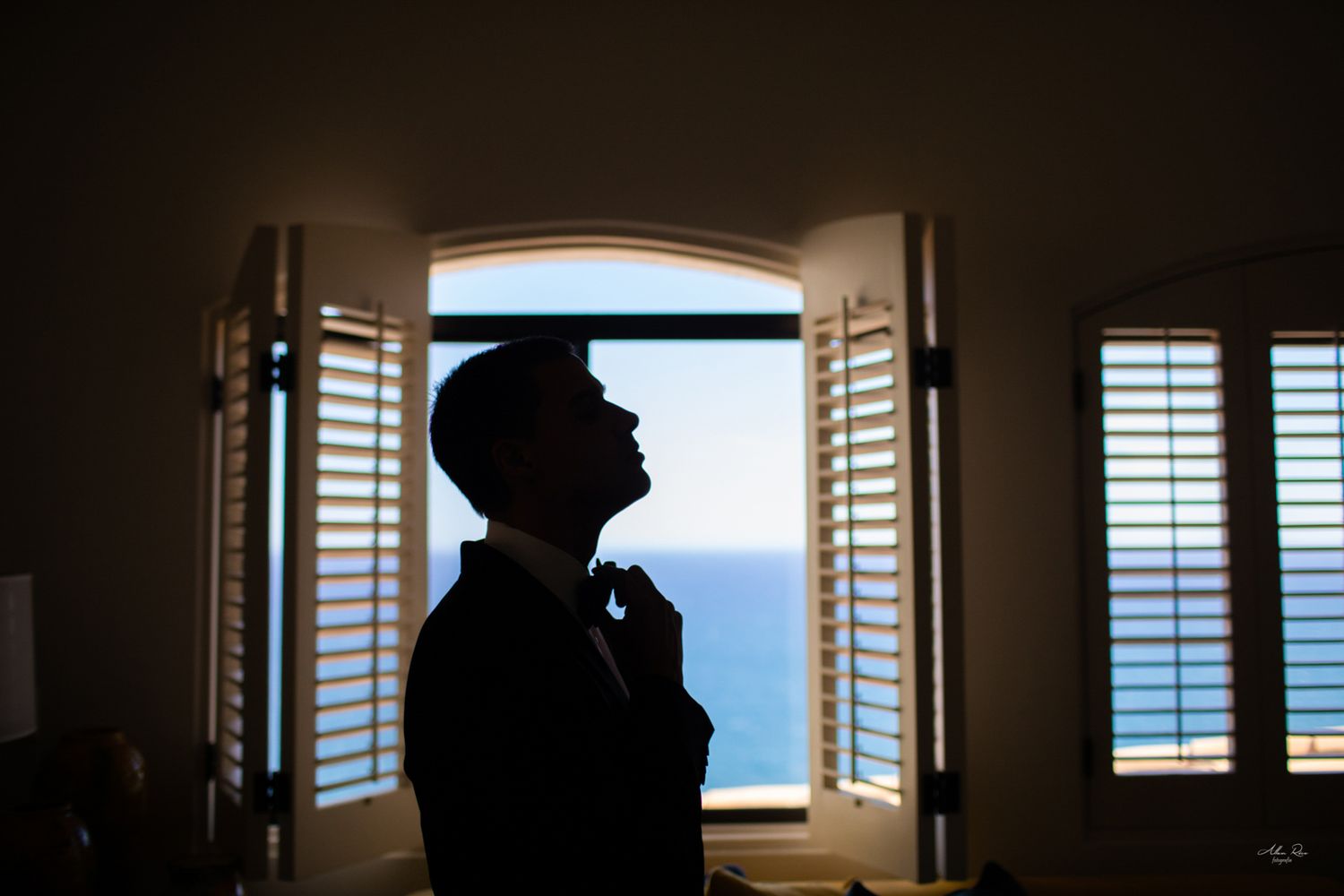 Groom silhouette adjusting bow tie by arched window with ocean view on wedding day.