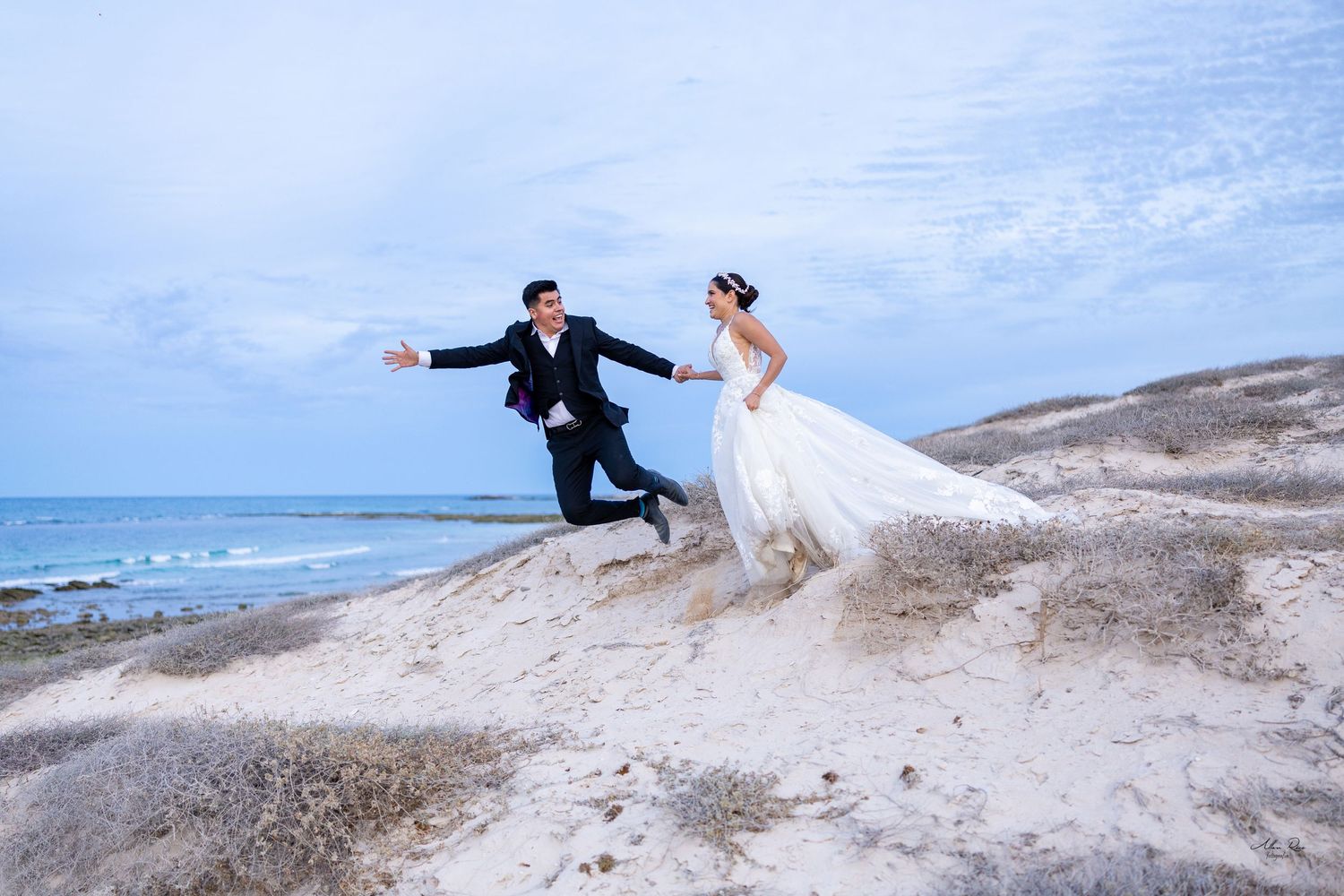 Joyful bride and groom jumping on sandy beach dunes with ocean and cloudy sky backdrop.