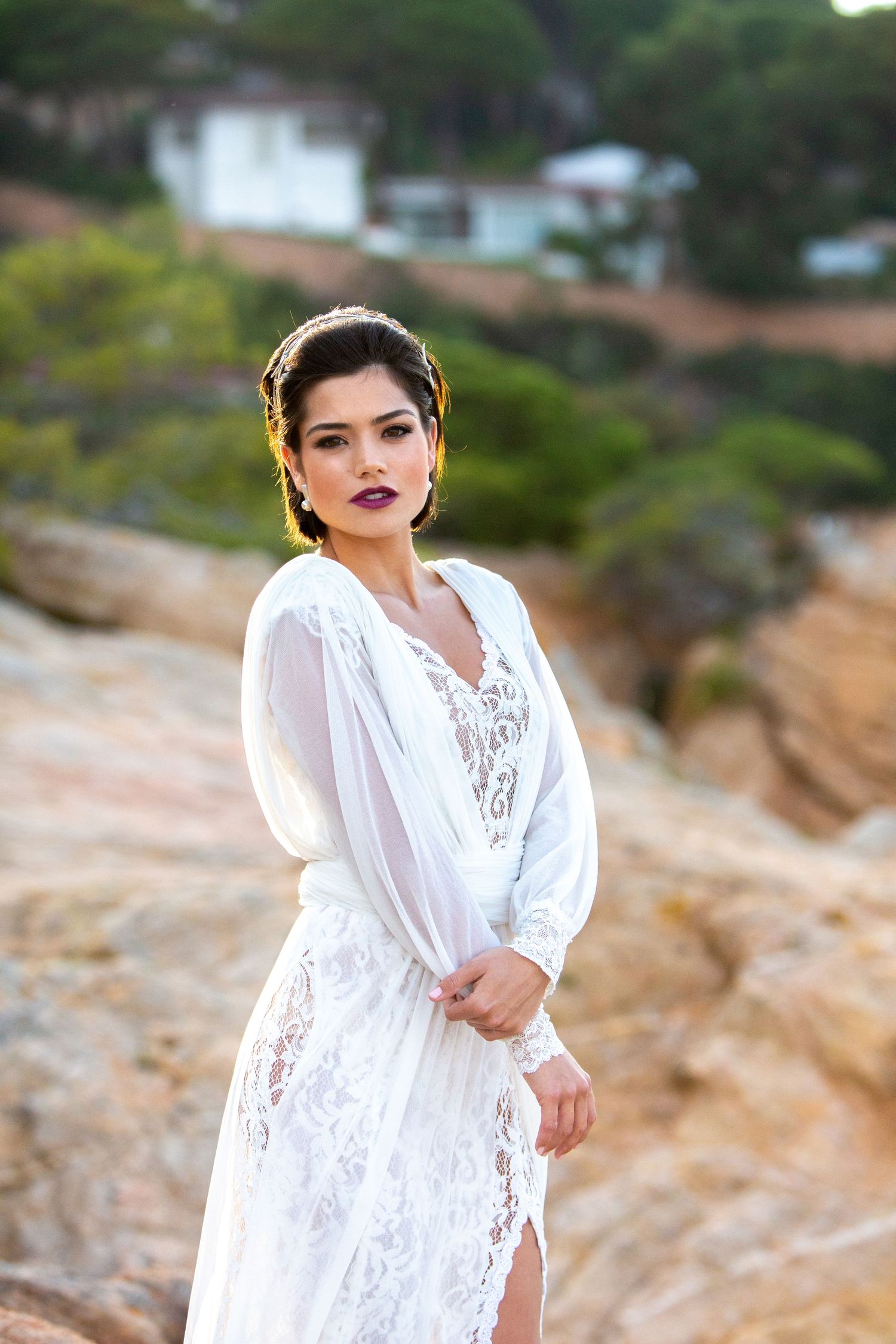 Bride in white lace long-sleeve wedding dress poses on rocky terrain with bold makeup and gold crown.