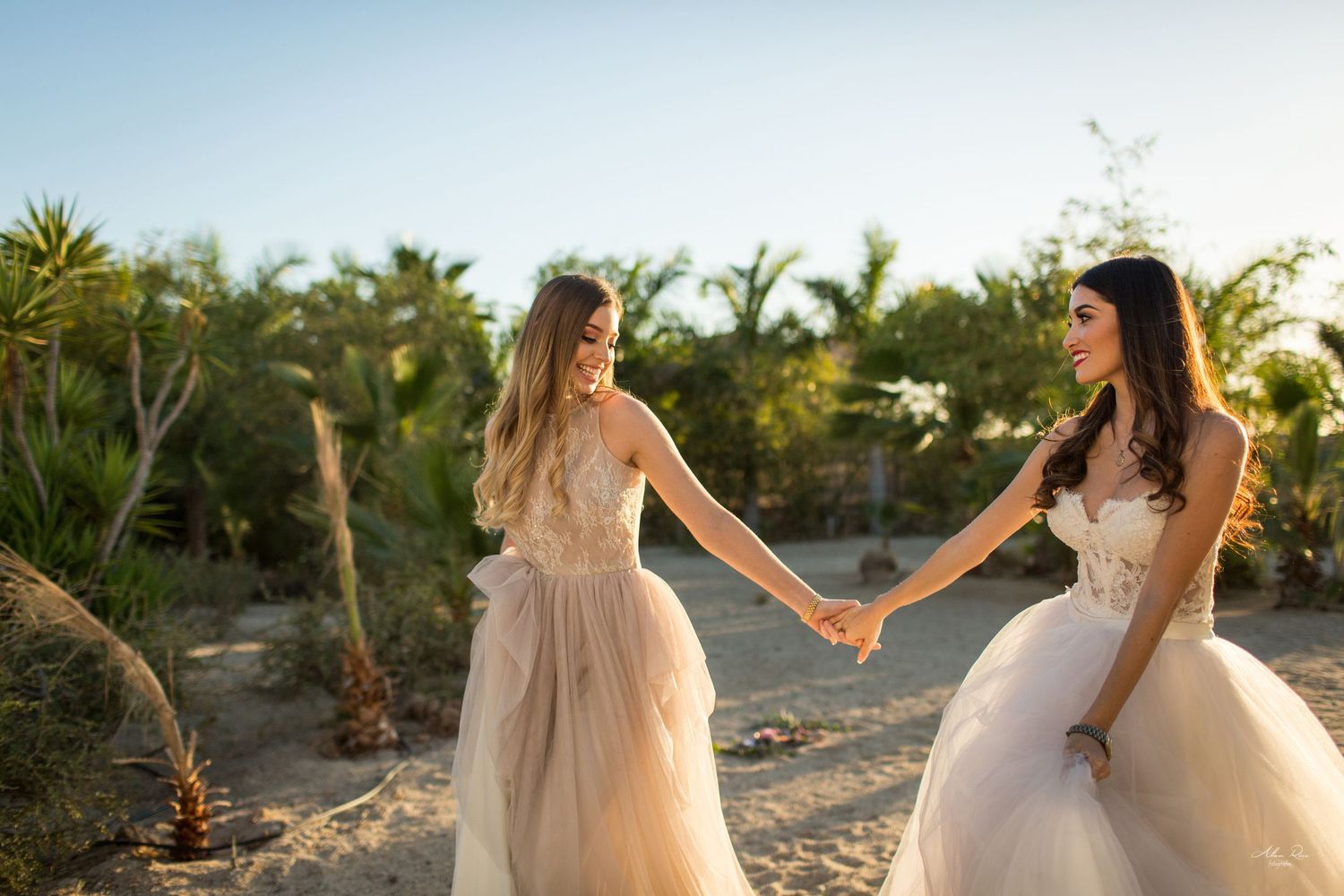 Two brides in flowing wedding gowns hold hands and smile while walking through a desert garden setting.
