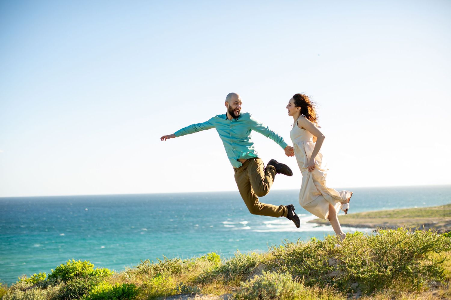 Joyful couple jumping together on a cliff overlooking turquoise ocean.