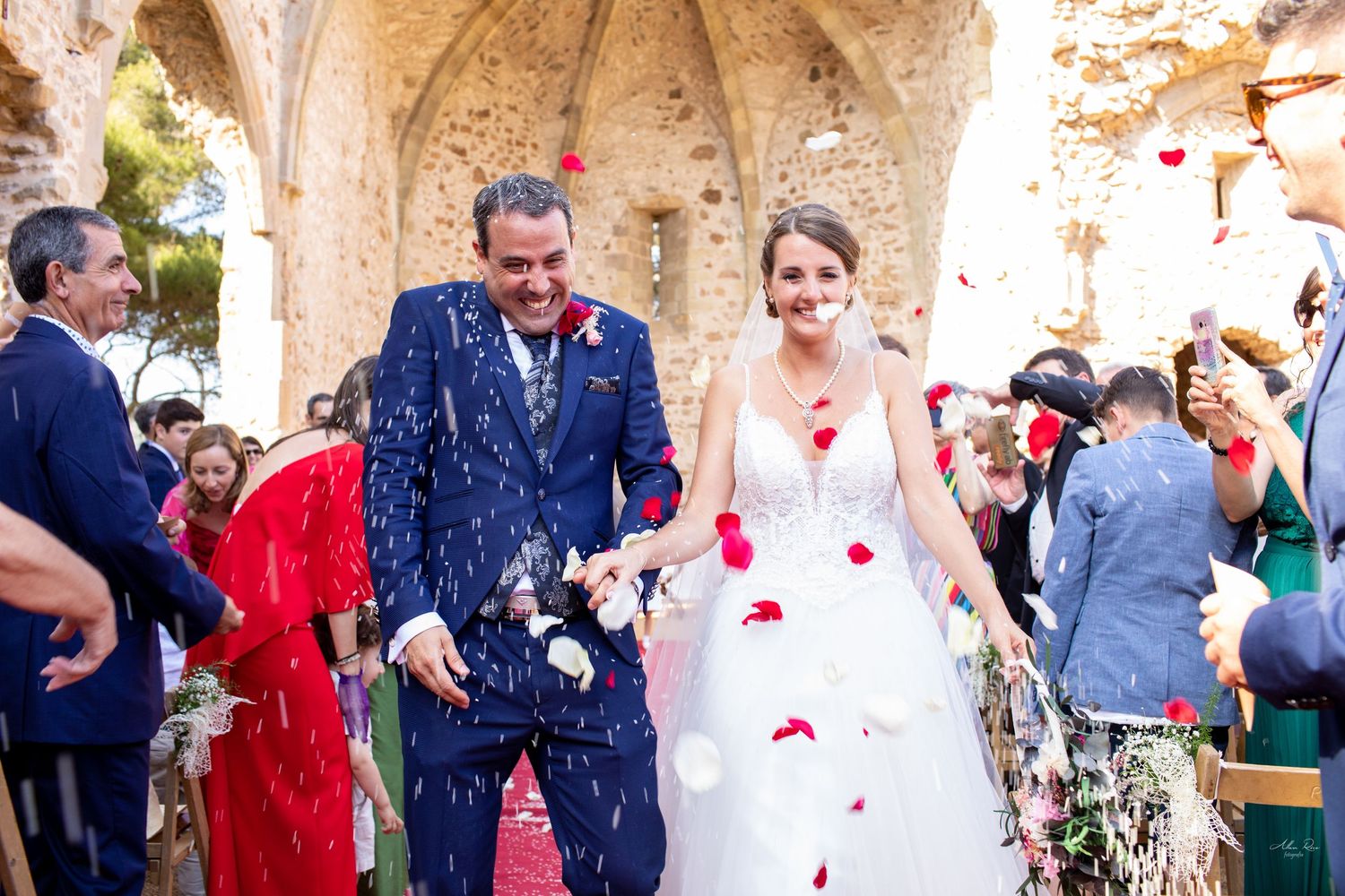 Newlyweds walk down the aisle as guests shower them with colorful rose petals in historic stone ruins.