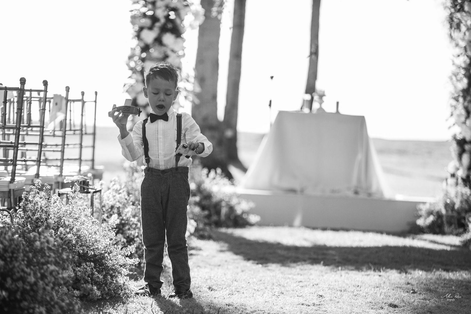 Young ring bearer in suspenders and bow tie stands in black and white outdoor wedding setting.