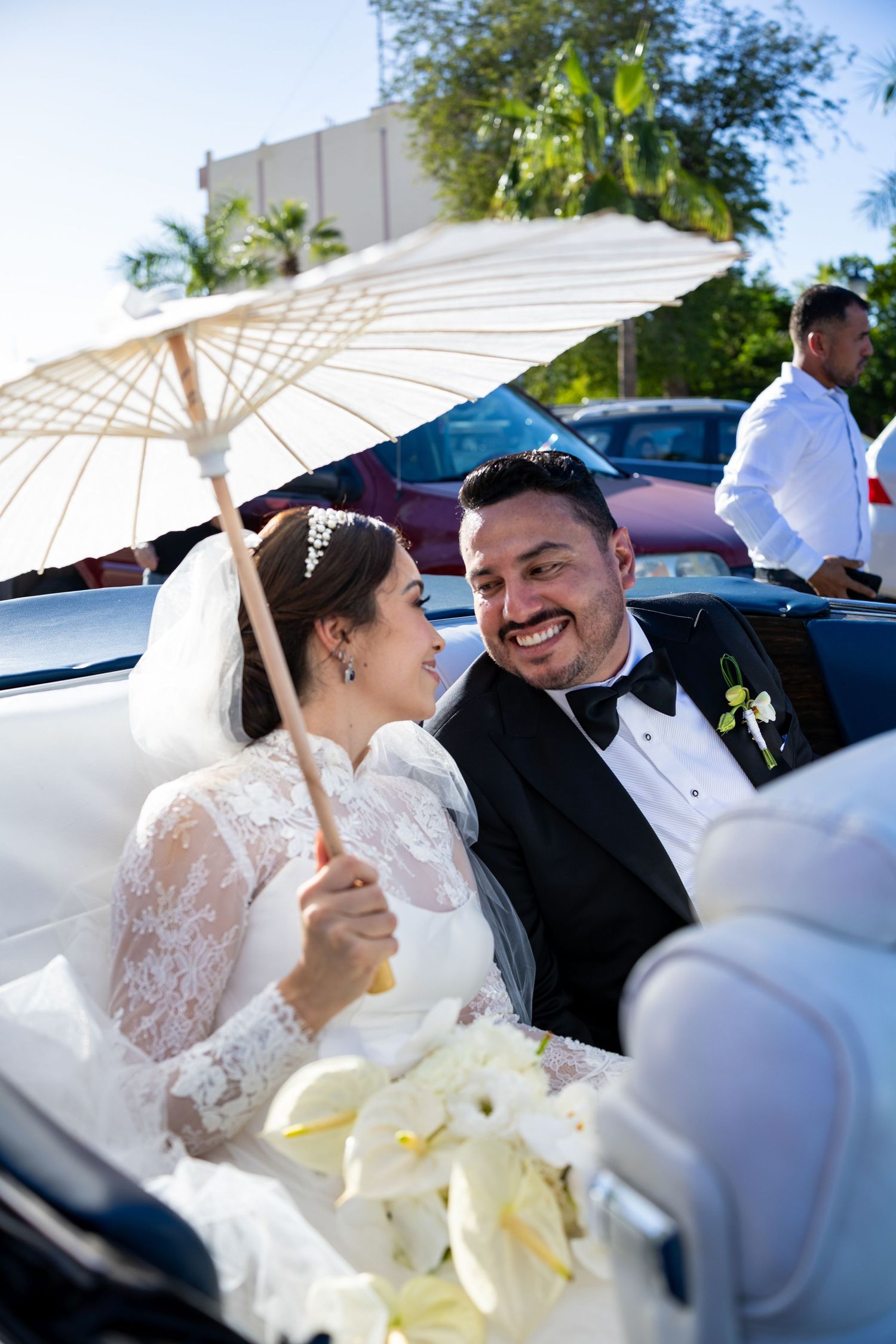 Bride and groom smiling together in vintage convertible with white parasol.