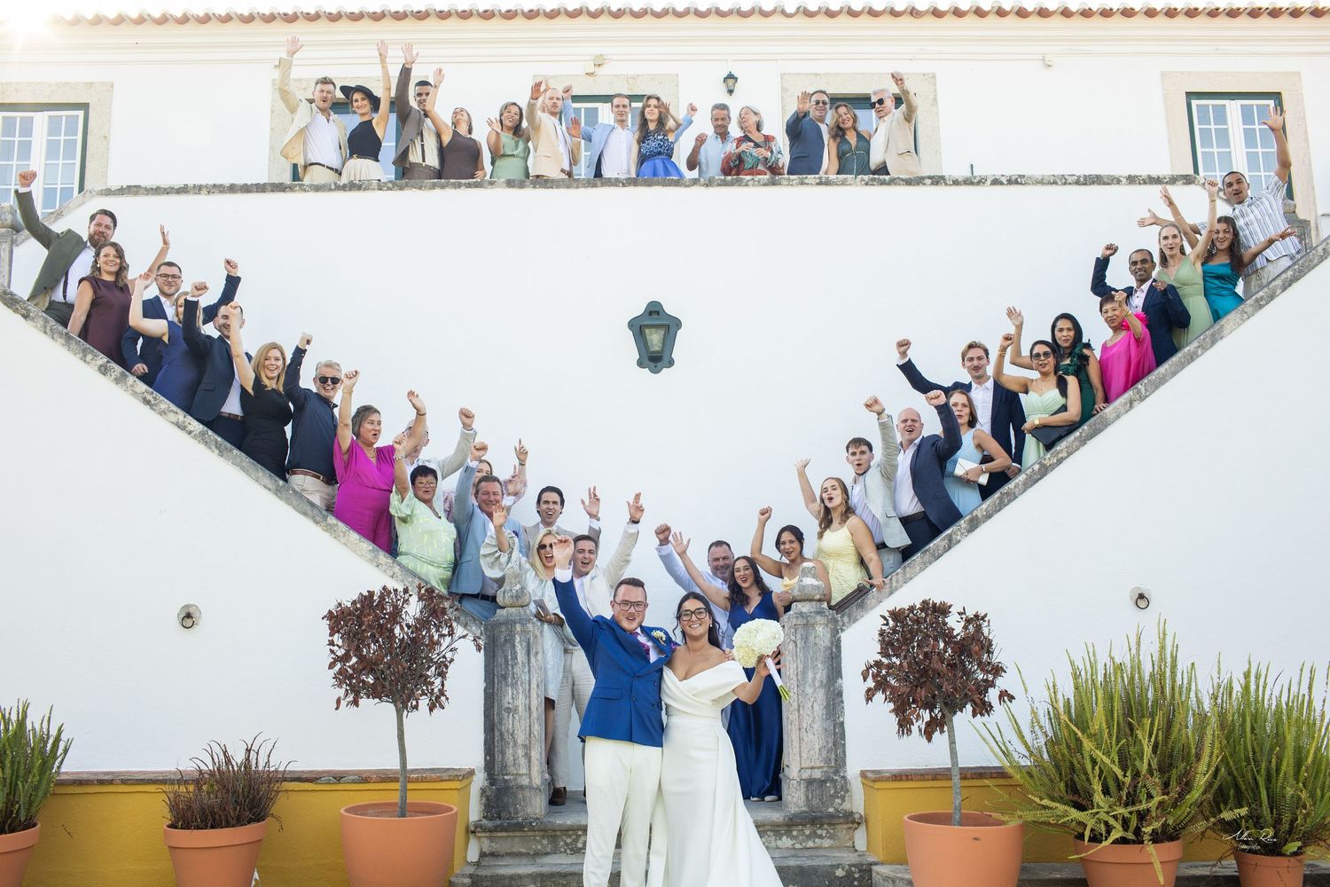Wedding party and guests pose on grand staircase celebrating with newlyweds at elegant venue.