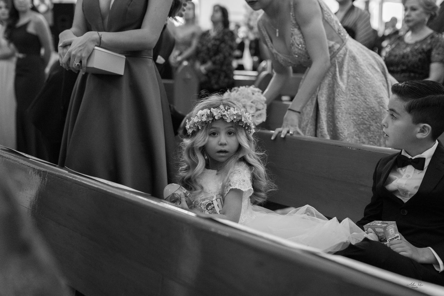 Adorable flower girl with floral crown sits in church pew during wedding ceremony.
