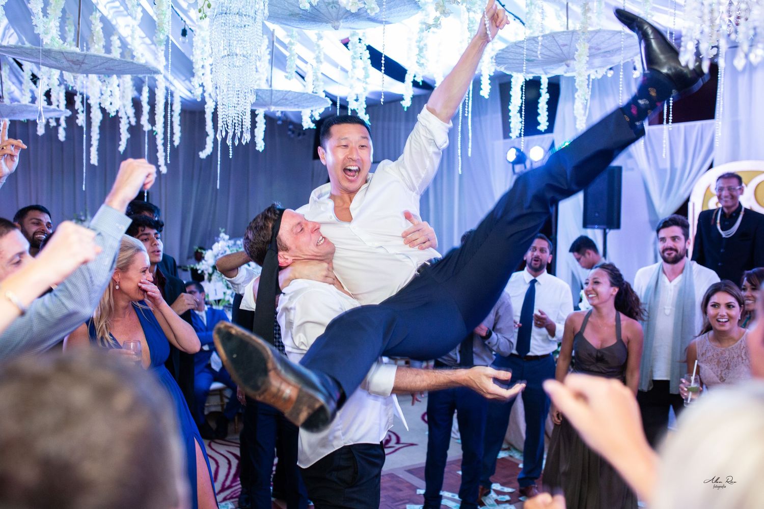 Guests lift a smiling man high in the air during a joyful wedding reception celebration on the dance floor.