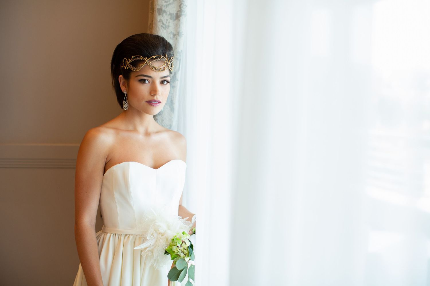 Elegant bride in strapless ivory gown with gold headpiece holds a bouquet while gazing out a window.
