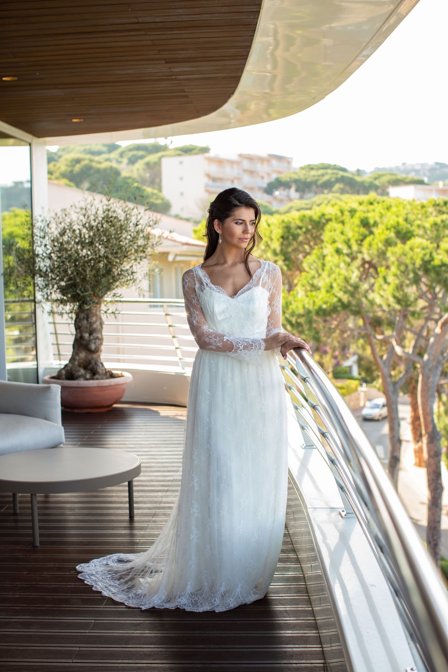 Bride in a long-sleeve lace wedding gown poses gracefully on a wooden balcony overlooking a sunlit Mediterranean landscape.
