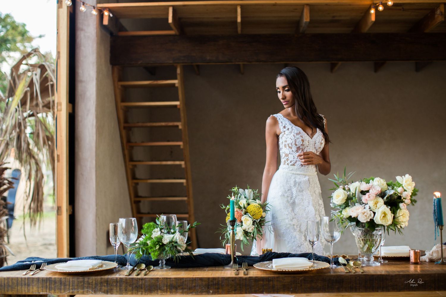 Bride in lace gown holding glass standing at elegantly set outdoor reception table with flowers.