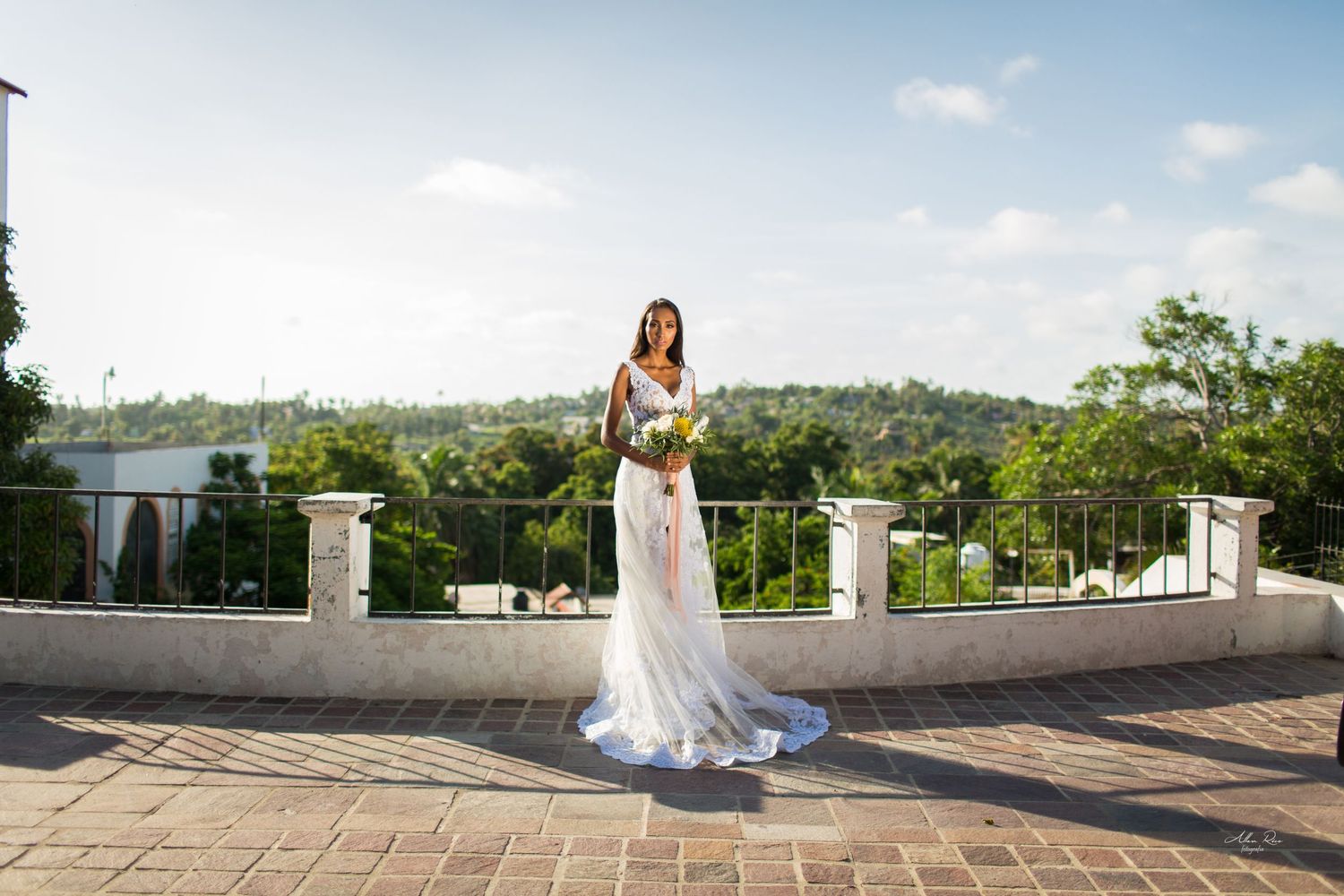 Bride in white lace gown holding bouquet stands on stone terrace with lush green landscape behind.