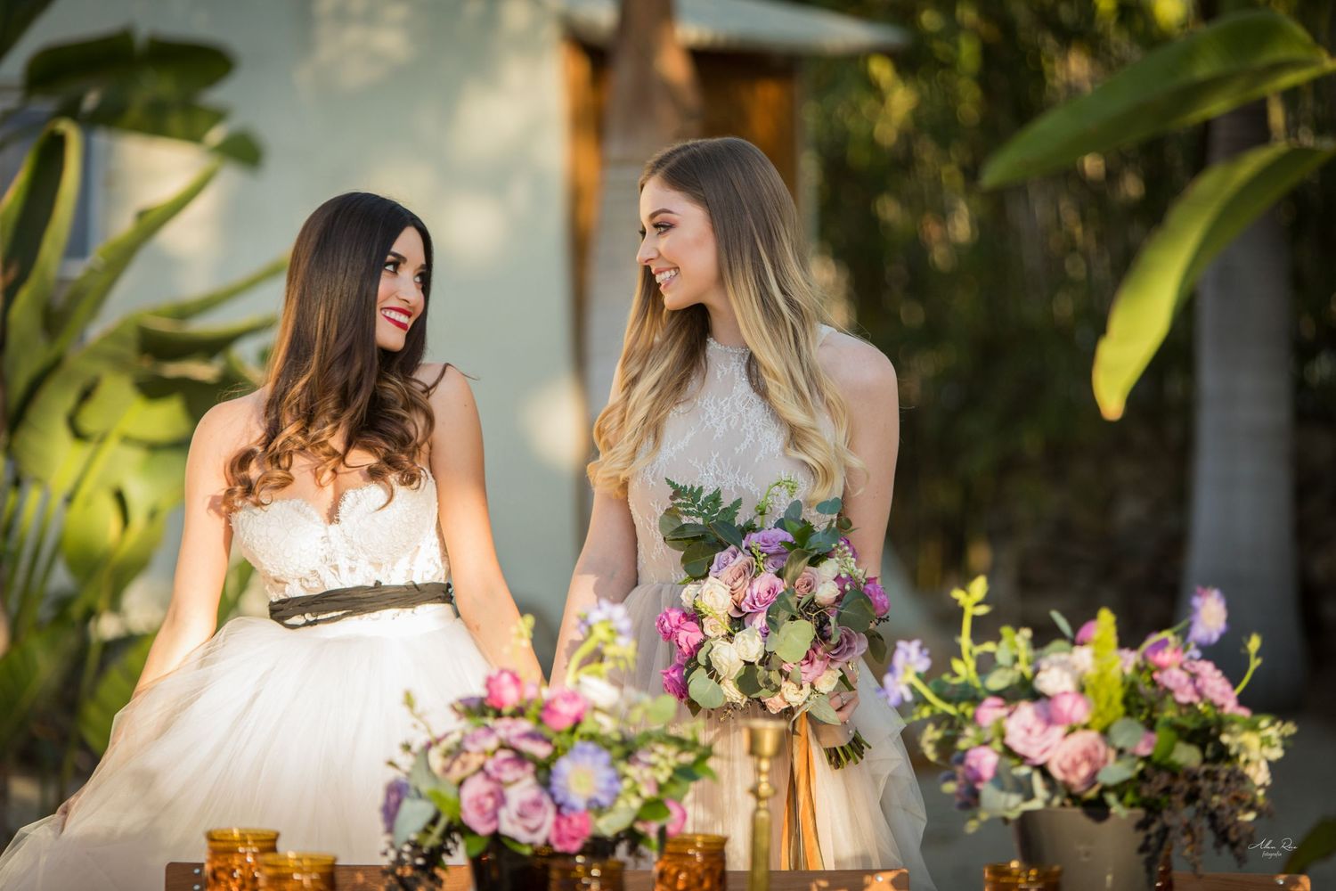 Two brides in white gowns smiling together beside a floral table arrangement outdoors.