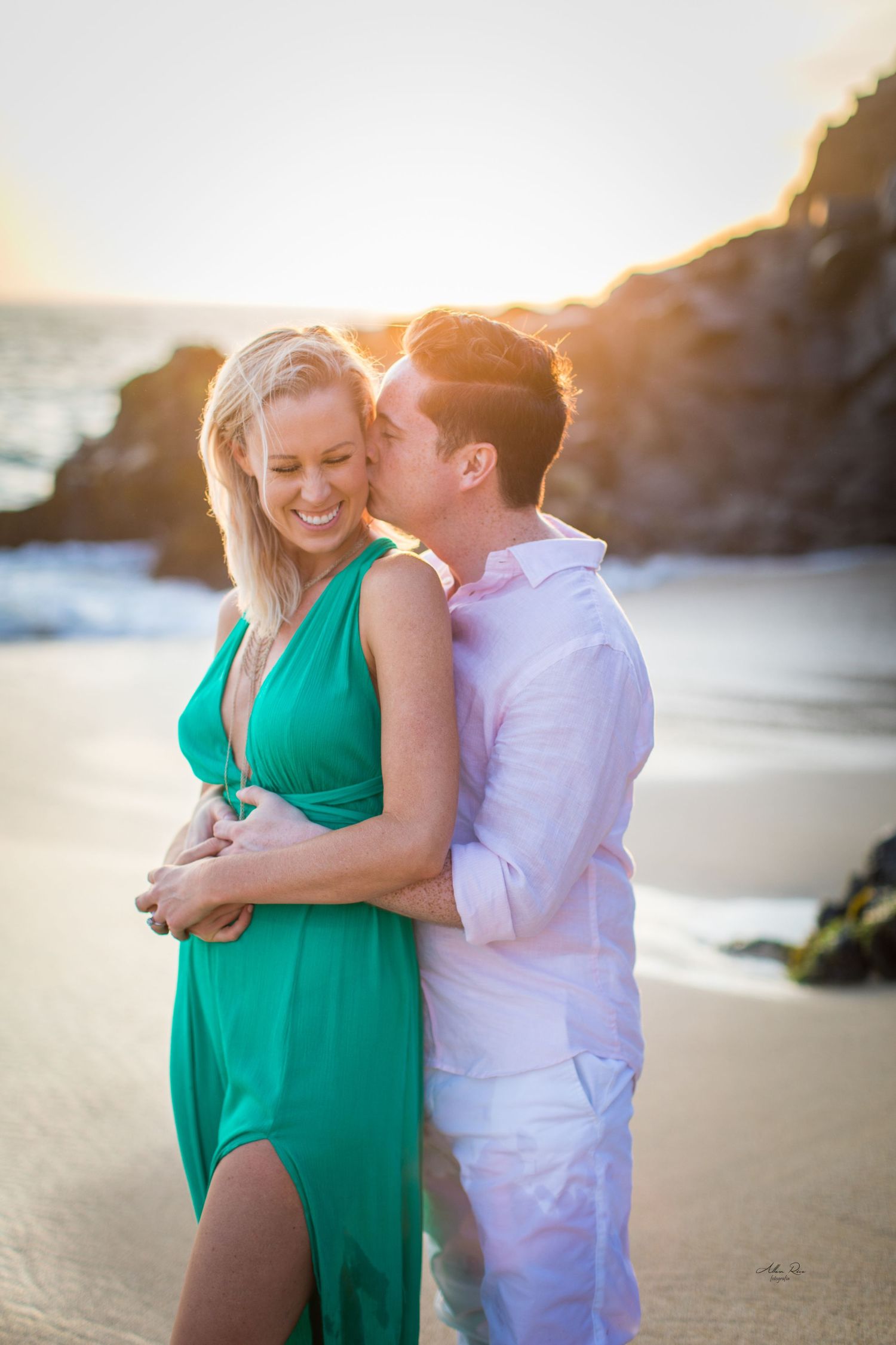 Man kissing woman on cheek as she laughs on a beach at sunset, woman wearing elegant green dress.