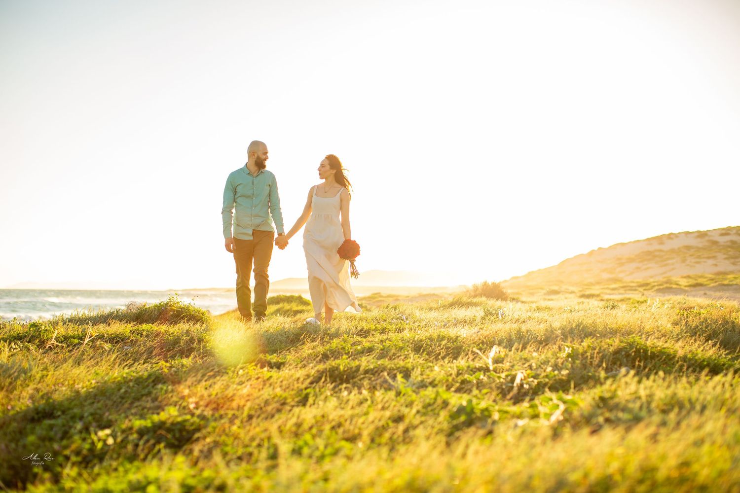 Couple holding hands walking through golden coastal meadow at sunset.