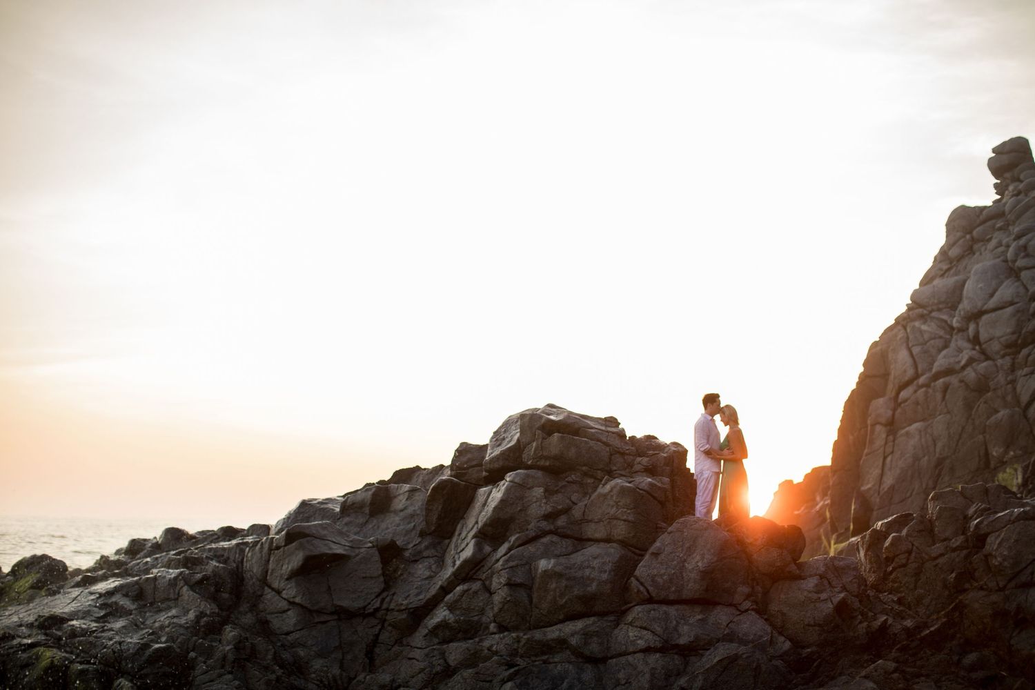 Romantic couple standing together on dramatic rocky cliffs silhouetted against a glowing golden sunset.