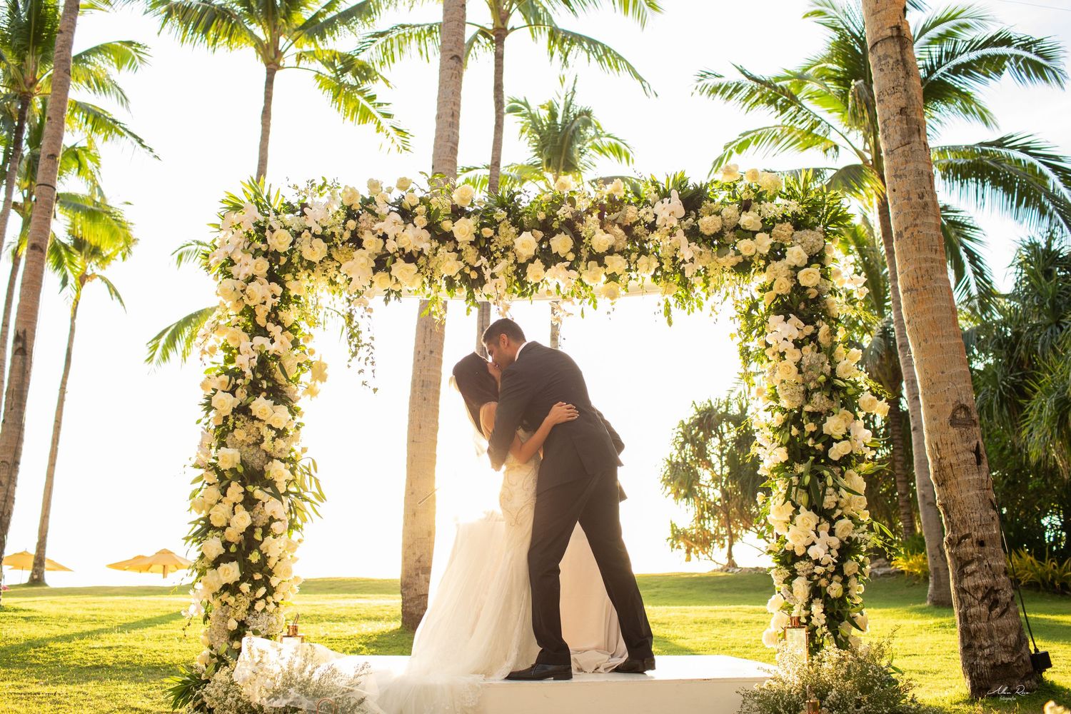 Bride and groom sharing a first kiss under a gorgeous white floral arch at a tropical outdoor wedding.