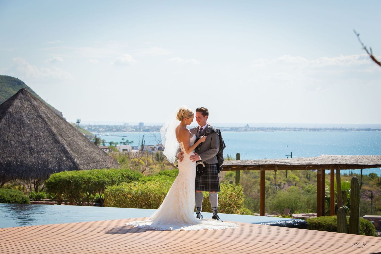 Bride and groom kissing on a wooden deck overlooking the ocean, groom wearing a traditional Scottish kilt.