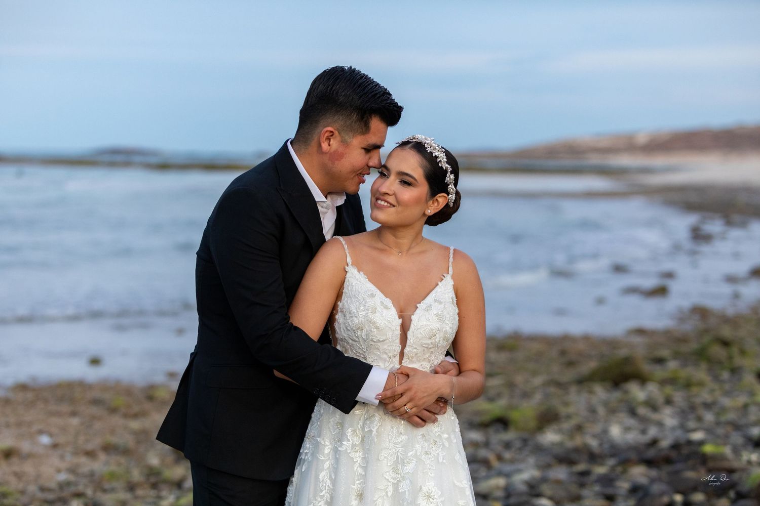 Groom tenderly embraces bride in lace wedding dress on rocky shoreline with ocean waves in the background.